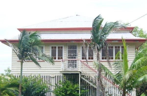 A raised wooden house with a metal roof, red trim, front steps with a railing, and surrounded by palm trees and greenery.